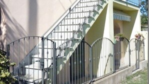 Un edificio de dos pisos con un balcón en el segundo piso, una escalera con barandillas de metal y un pequeño porche con plantas en macetas.