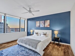 A bedroom with a large bed, two bedside tables with lamps, a ceiling fan, and a view of buildings through a large window. - Luxury Apt w/Parking CityView/Pool/Gym/PoolTable (Dallas)