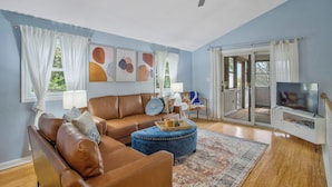 A living room with a brown leather sofa, a blue ottoman, a television on a white stand, and a ceiling fan.