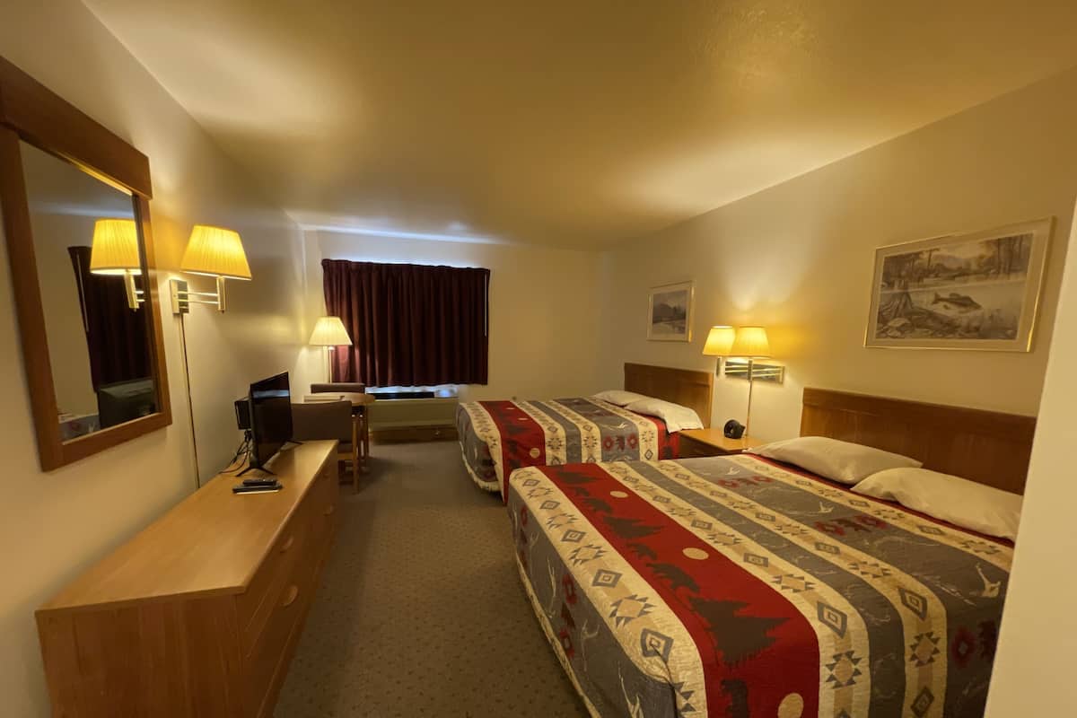 A hotel room with two beds, a desk, a chair, a TV, and lamps.