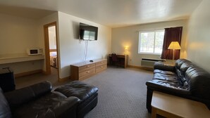A living room with a flat-screen TV, a desk with a chair, a sofa, and a lamp. - AuSable Valley Inn (Mio)