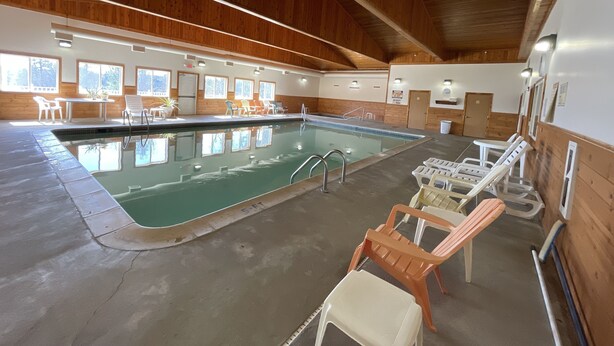 An indoor swimming pool with a wooden ceiling, clear glass walls, and a sunlit room with chairs and tables. - AuSable Valley Inn (Mio)