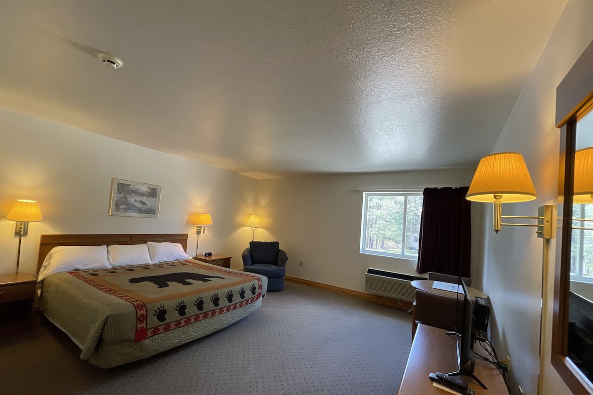 A hotel room with a bed, two bedside lamps, a chair, a desk with a computer, and a window with curtains.