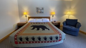 A hotel room with a bed, two bedside lamps, a chair, and a painting on the wall. - AuSable Valley Inn (Mio)
