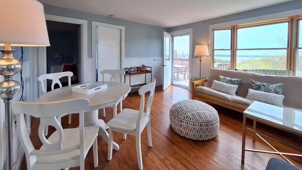 A spacious living area with a dining table and chairs, a sofa, a patterned ottoman, and large windows offering a view of the outdoors. - Attached Apartment Near It All. Organic Farmstead. Stunning Views. Pet-friendly. (New Shoreham)