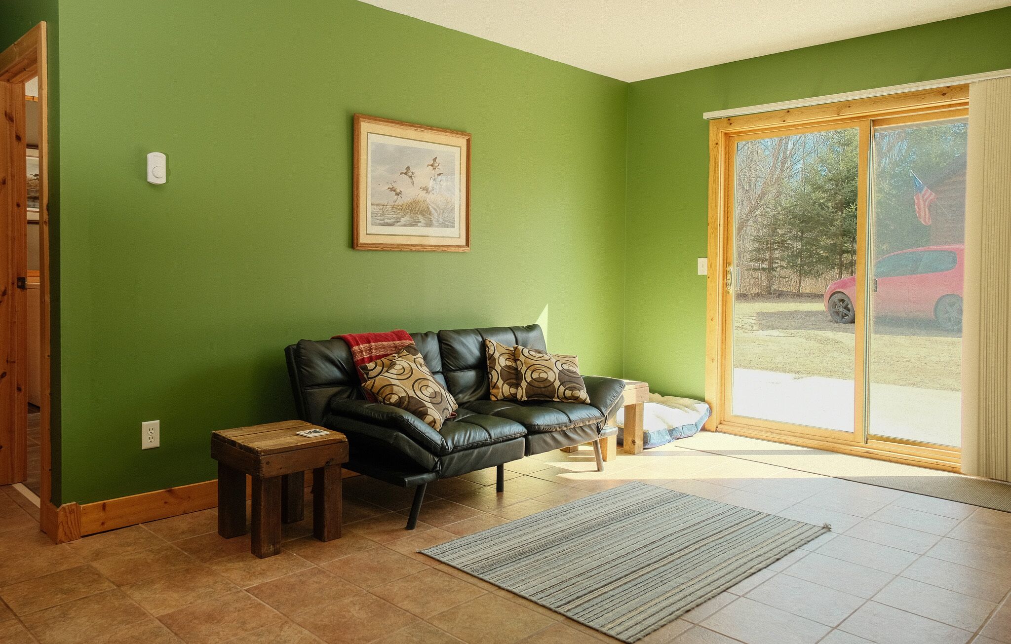 A living room with a green wall, a black leather sofa, a small wooden table, a framed picture, and a sliding glass door leading outside.