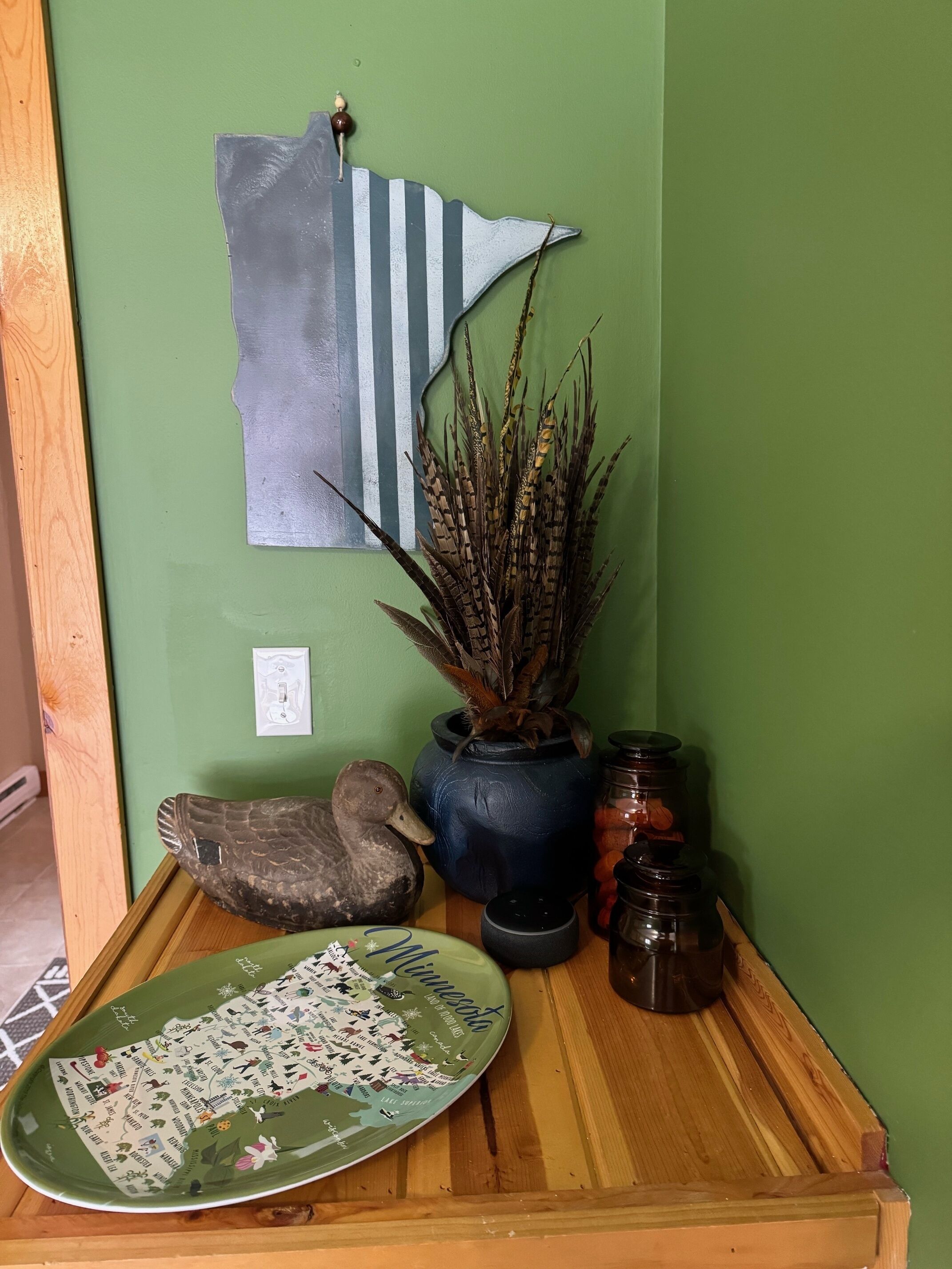 A wooden table with a decorative plate, a duck sculpture, a vase with dried plants, a blue pot, and a wall-mounted flag.