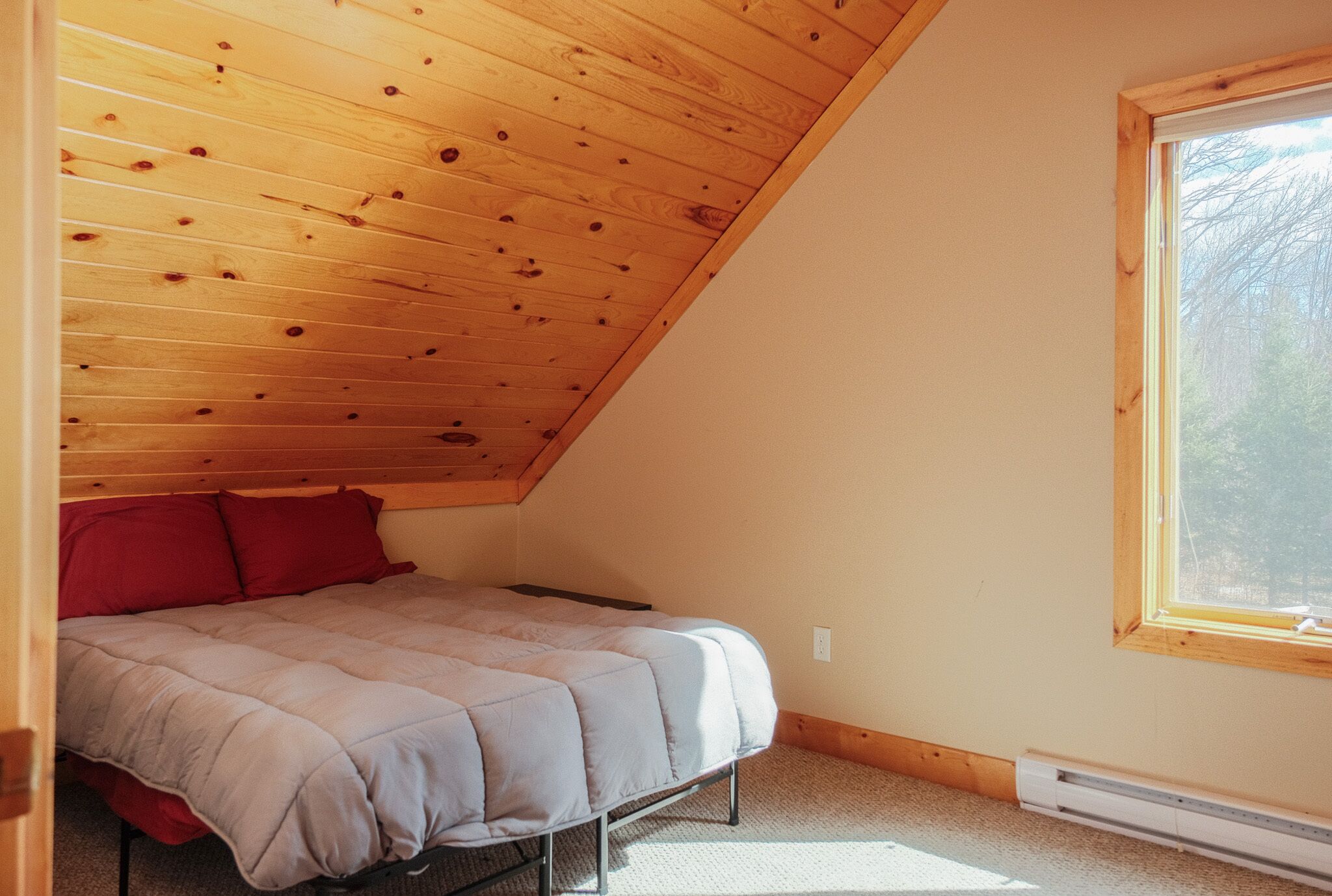 A bedroom with a sloped ceiling, a bed with a grey duvet, red pillows, and a window with a view of trees.