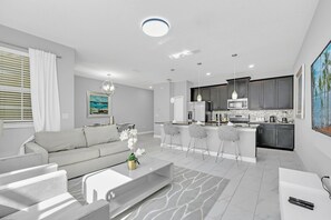 A modern living room with a sofa, coffee table, and a dining area with bar stools. The kitchen features dark cabinets and a central island.