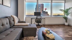 A modern living room with a grey sofa, a small table with flowers, and a view of a cityscape through large windows. - Luxury Apt In City Heart with Pool Gym & PoolTable (Dallas)