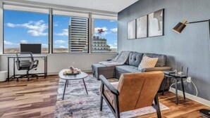 A modern living room with a large window, a ceiling fan, a grey sofa, a coffee table, and a desk with a chair. - Luxury Apt In City Heart with Pool Gym & PoolTable (Dallas)