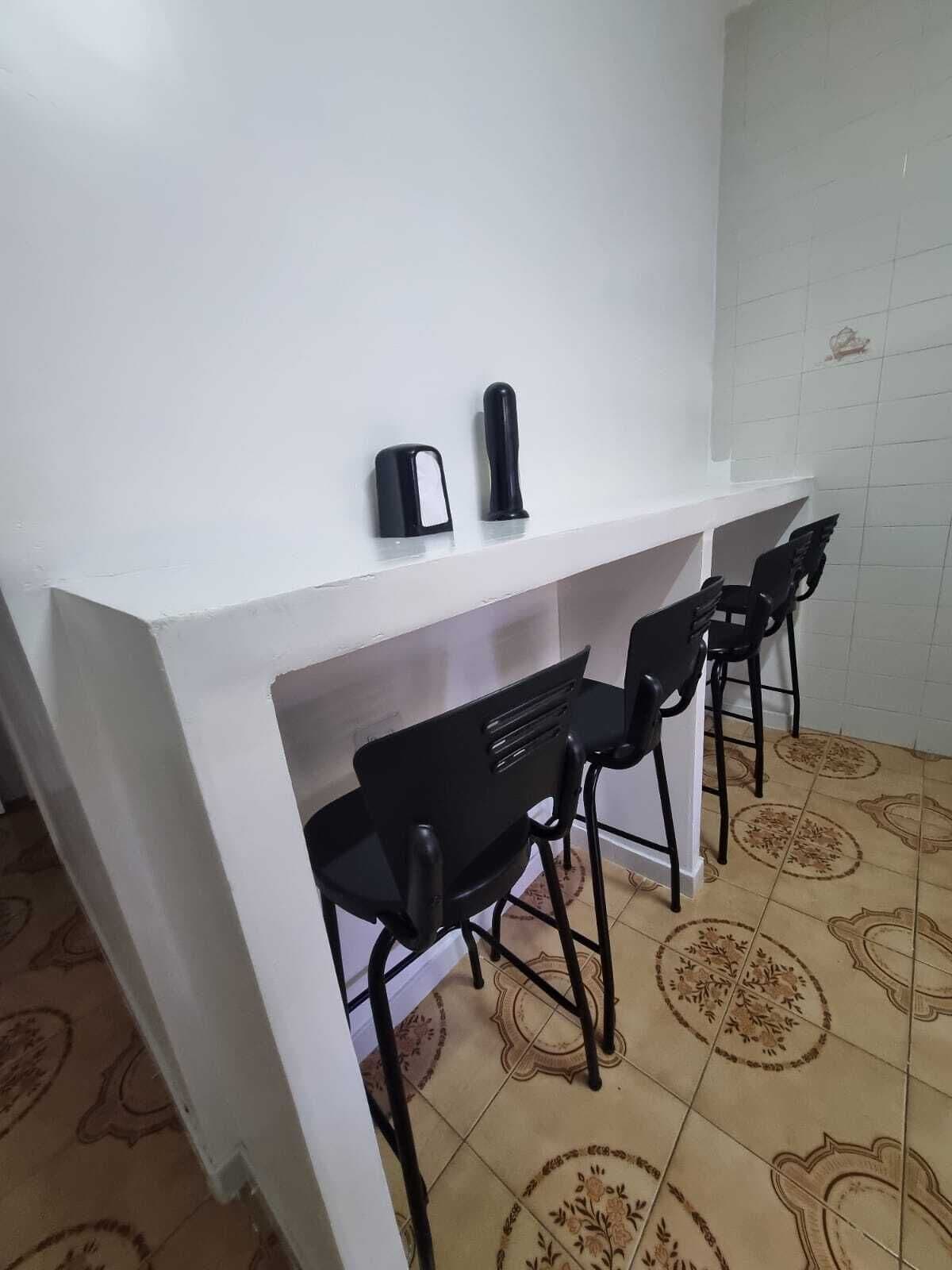A small, white kitchenette with a countertop, two black chairs, and a coffee maker.
