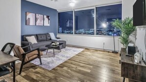 A modern living room with a large window, a sofa, a coffee table, and a TV on a wooden stand. - Luxury 1 Bhk Apt w/Parking Pool / Gym/ City View (Dallas)
