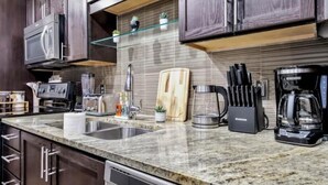 A modern kitchen with dark wood cabinets, a granite countertop, stainless steel appliances, and a glass shelf above the sink. - Luxury 1 Bhk Apt w/Parking Pool / Gym/ City View (Dallas)