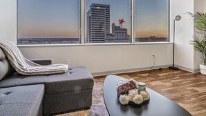 A modern living room with a grey sofa, a small table with decorative items, and a large window offering a city view. - Luxury 1 Bhk Apt w/Parking Pool / Gym/ City View (Dallas)