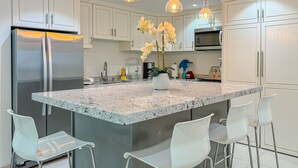 A modern kitchen with white cabinets, a stainless steel refrigerator, a granite island with bar stools, pendant lights, and a flower vase. - Downtown Oceana Condo/2 Min Walk to Papas&Beer (Playas de Rosarito)