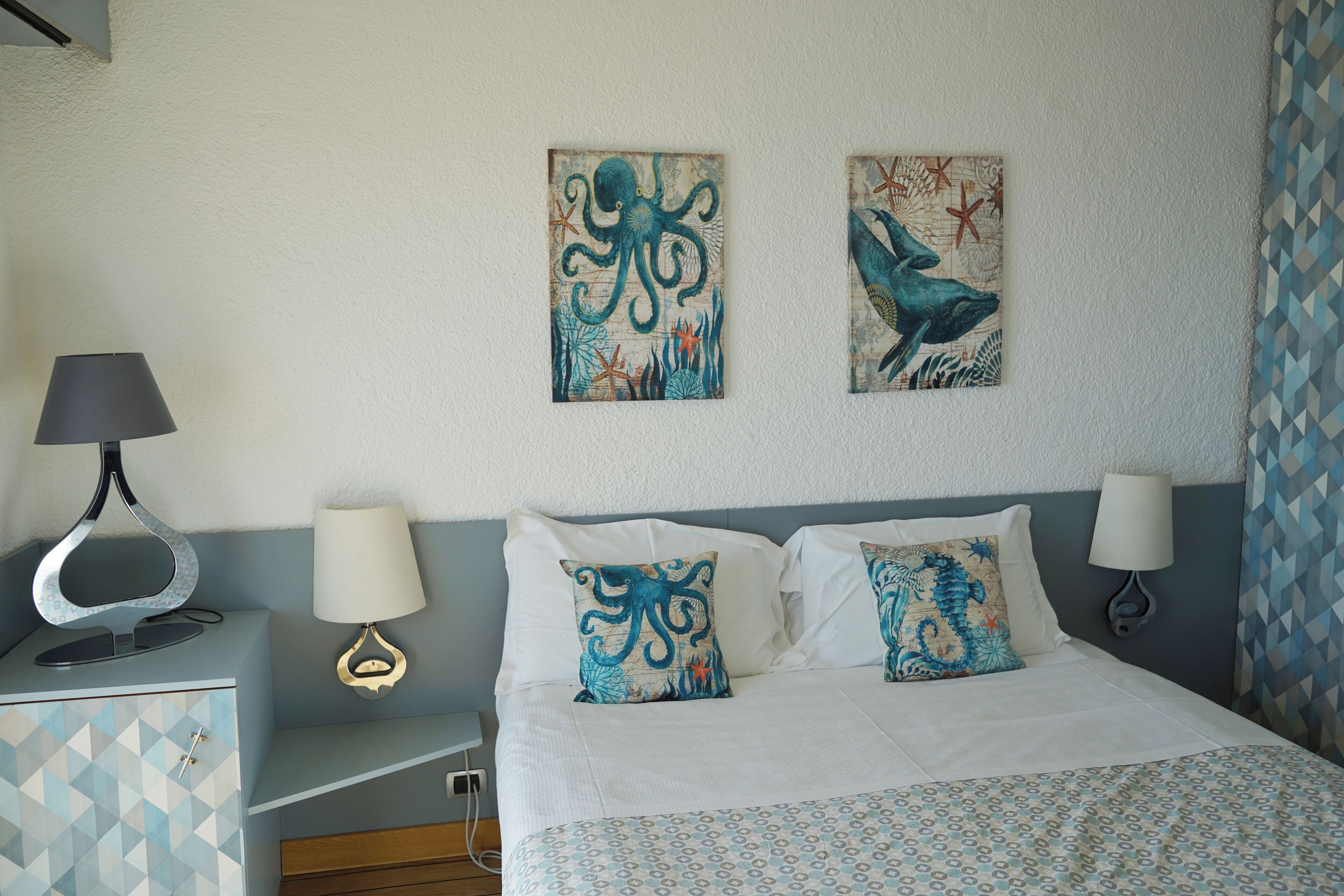 A neatly made bed with patterned bedding, two wall-mounted artworks depicting sea creatures, and a bedside table with a lamp.