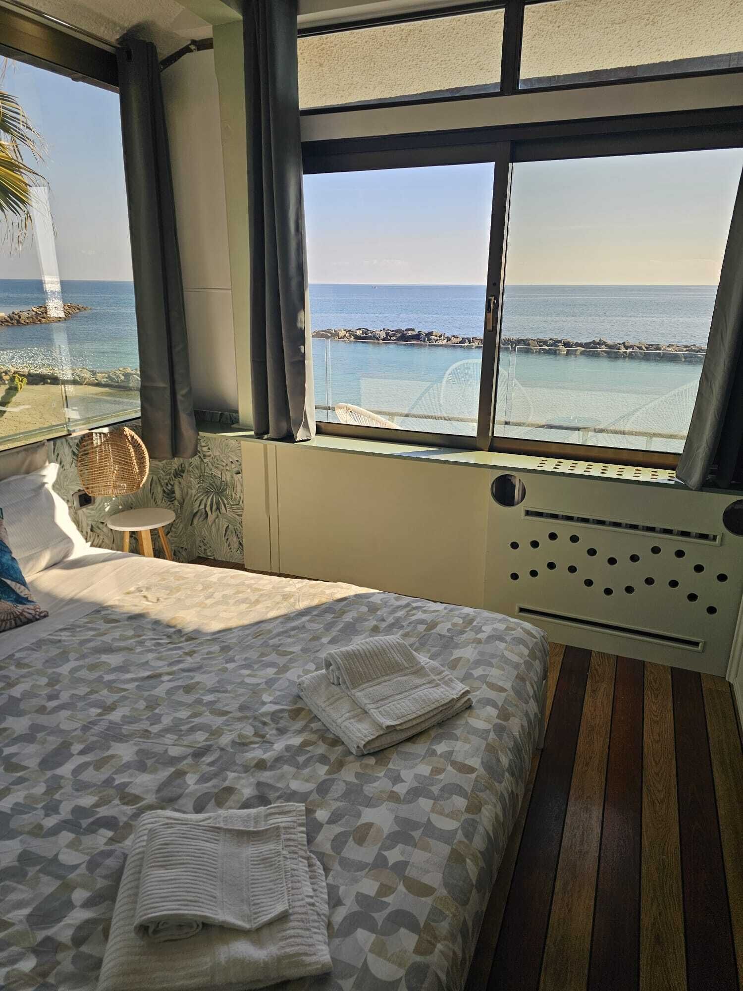 Una habitació d'hotel amb un llit, una taula petita, una cadira i un finestral gran que dona al mar i les roques.