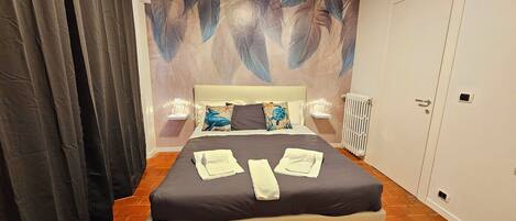 A neatly made bed with pillows and towels, a wall with a feather pattern, a radiator, and a door.