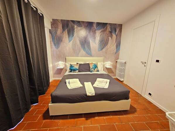 Un lit bien fait, avec des oreillers et des serviettes, un mur orné d’un motif de plumes, un radiateur et une porte.