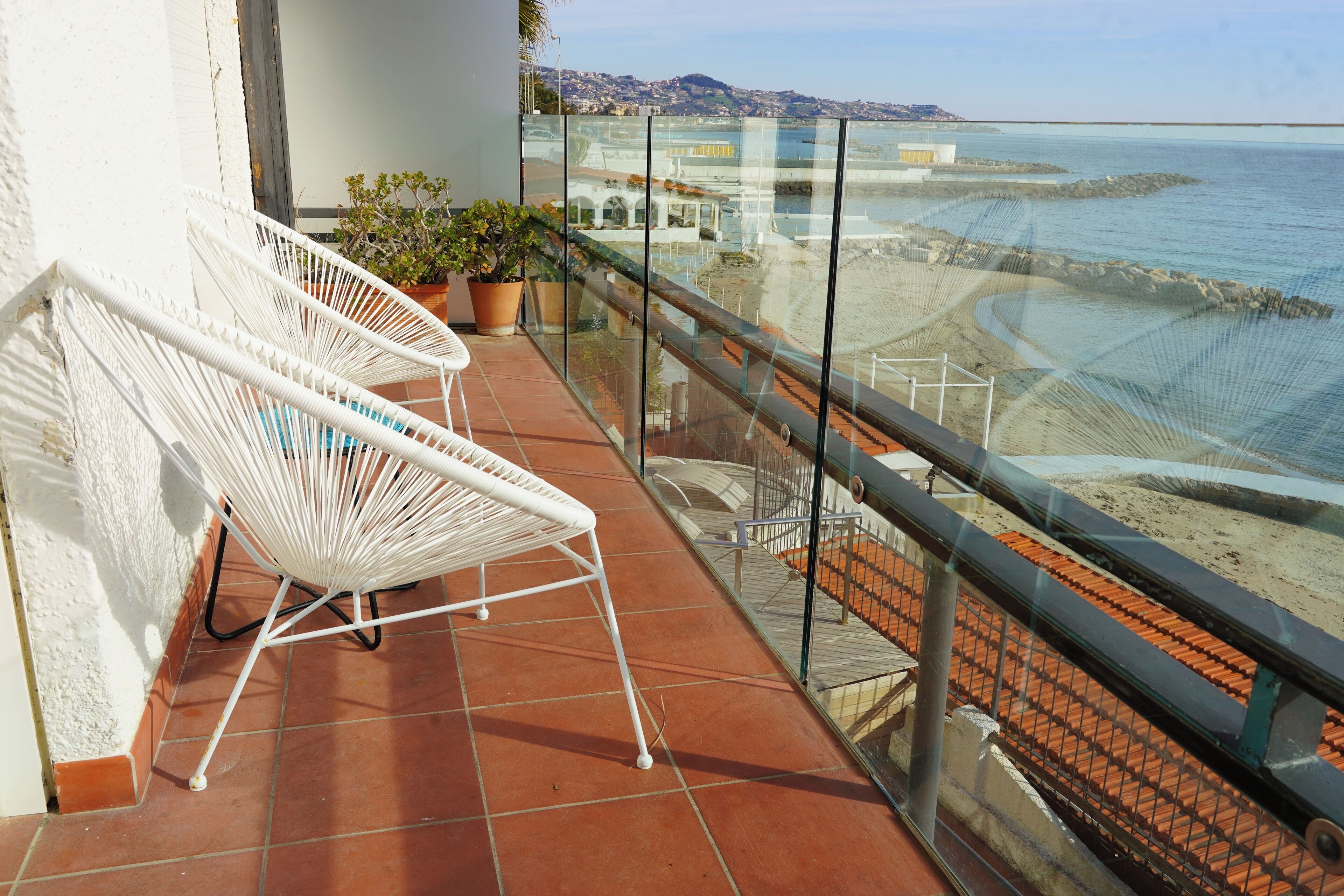 A balcony with white wicker chairs, potted plants, and a view of the sea and beach.