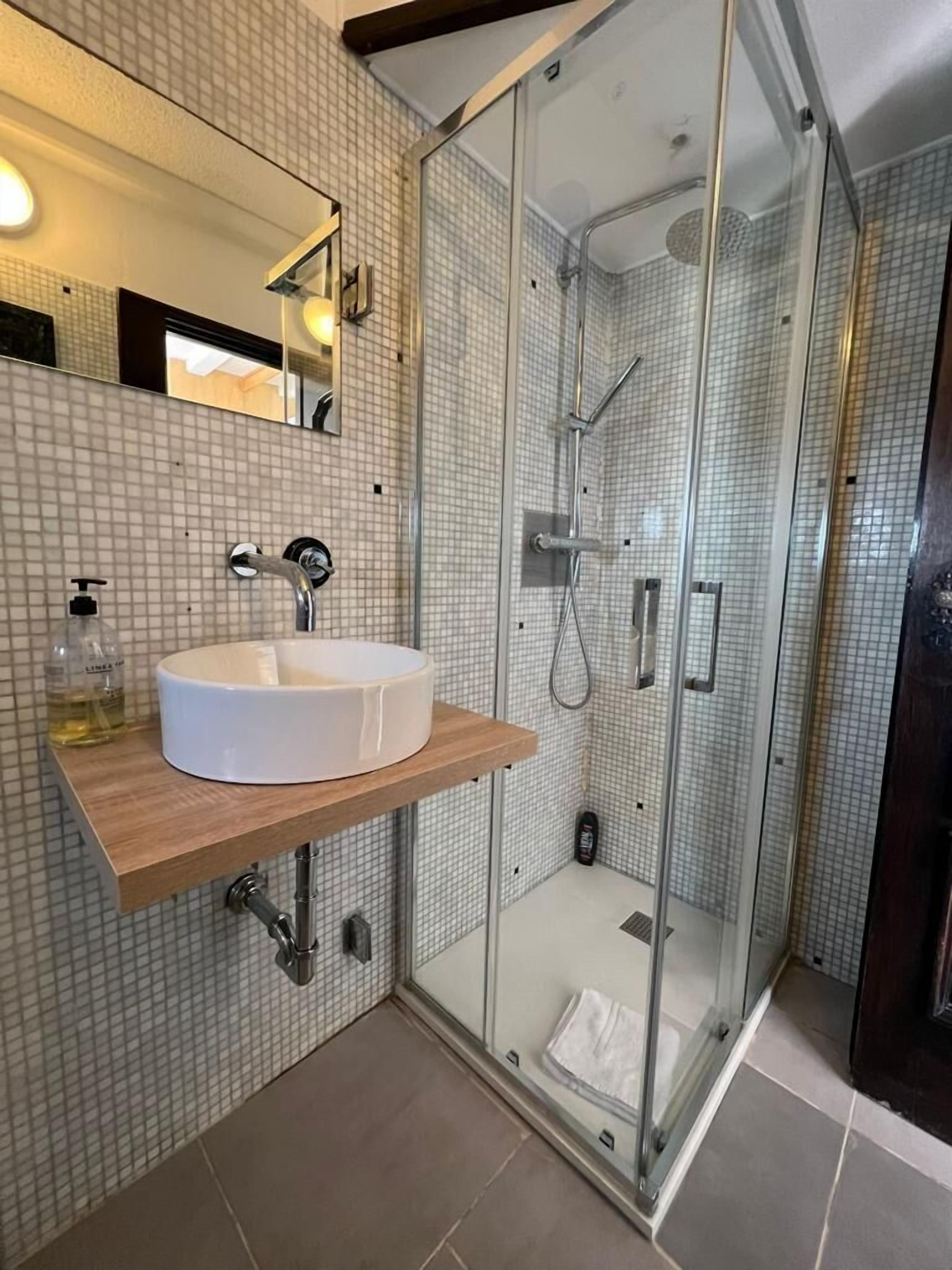 Un baño moderno con ducha envuelta en vidrio, un lavamanos blanco sobre encimera de madera y salpicadero de azulejos.
