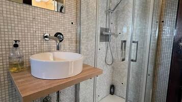 Un baño moderno con ducha envuelta en vidrio, un lavamanos blanco sobre encimera de madera y salpicadero de azulejos.