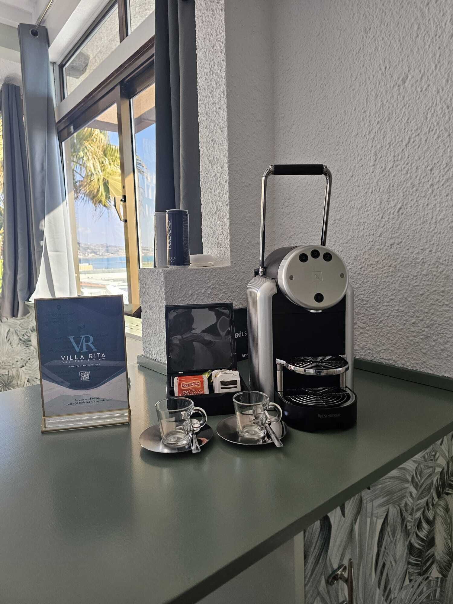 A minibar setup with a coffee machine, a box of Enjus coffee, and two glasses on a counter near a window with a view of palm trees.