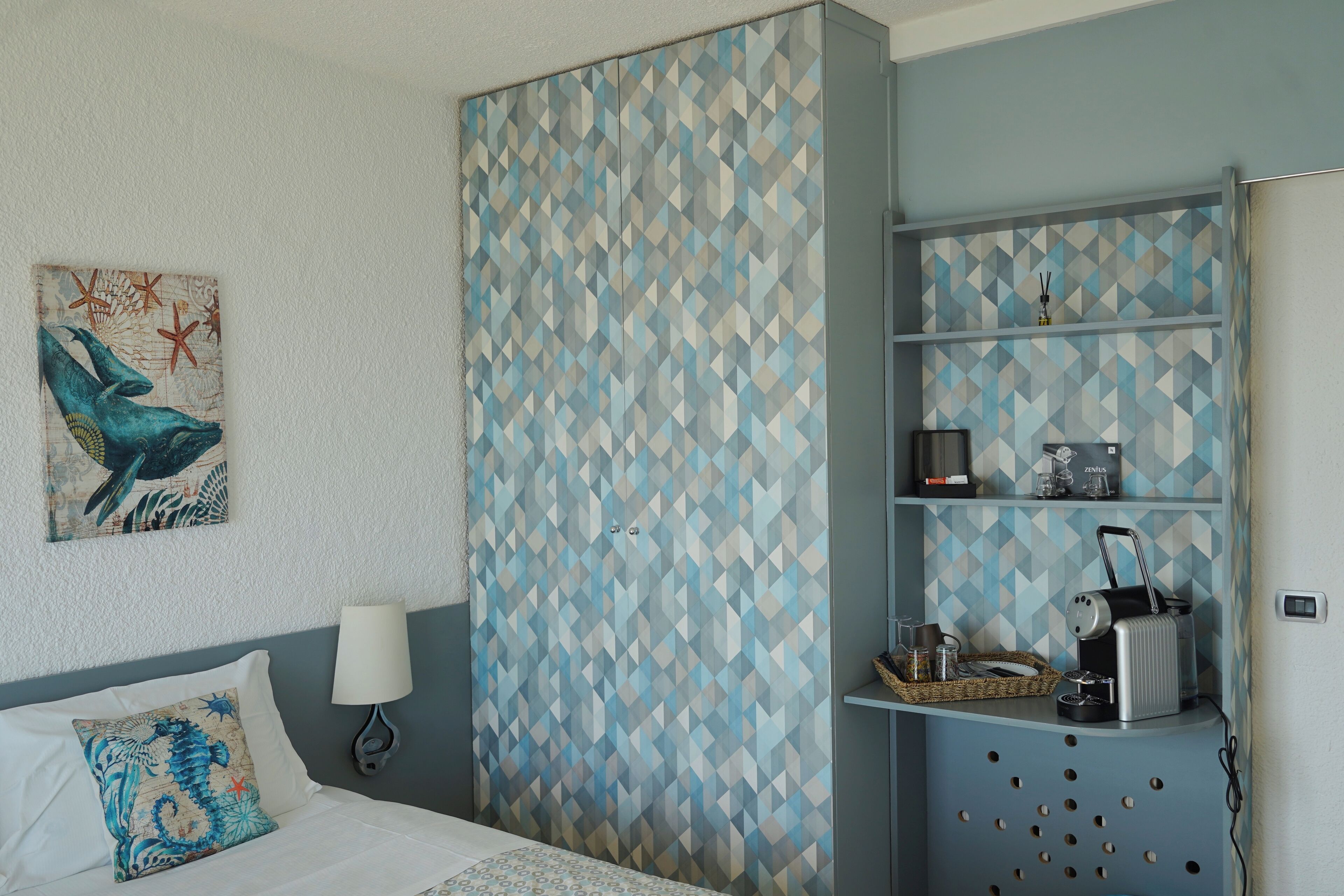 A bedroom with a bed, a nightstand, a lamp, a wall with a geometric pattern, and a shelf with various items.