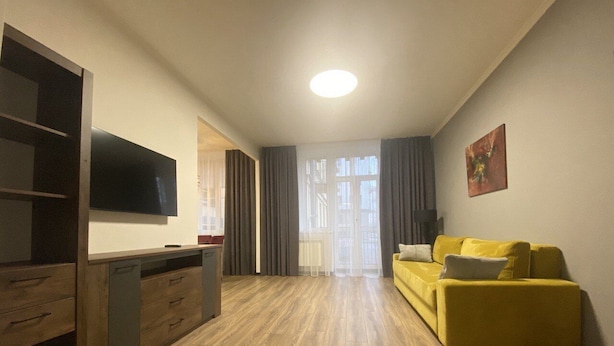 A modern living room with a wooden floor, a yellow sofa, a flat-screen TV, and a sliding glass door leading to a balcony. - Apartment  Home Apart Minsk Zaharova 24 (Minsk)