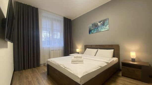 A hotel room with a large bed, a bedside table with a lamp, a painting on the wall, and a window with curtains. - Apartment  Home Apart Minsk Zaharova 24 (Minsk)