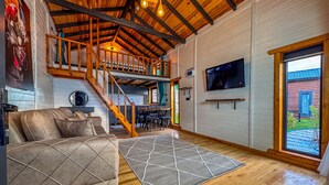 A spacious living area with a high ceiling, wooden beams, a sofa, a rug, a TV, and a staircase leading to an upper level. - AYVALIK TRİNGO BUNGALOV (Ayvalik)