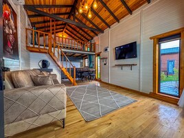 Uma área de estar espaçosa com teto alto, vigas de madeira, um sofá, um tapete, uma TV e uma escada que leva a um andar superior.
