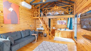 A cozy cabin interior with a sofa, a dining area, a staircase, and a large window. - AYVALIK TRİNGO BUNGALOV (Ayvalik)