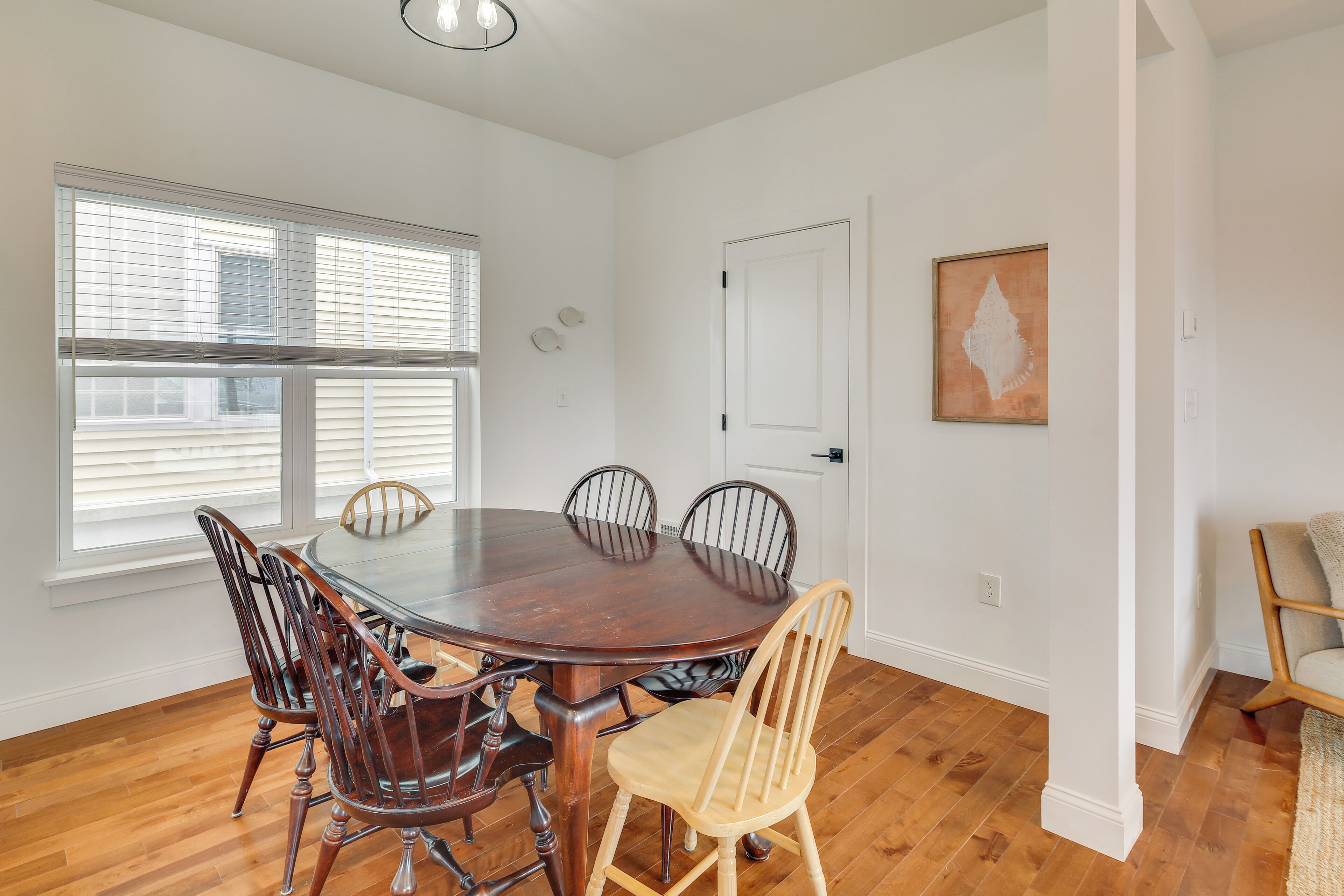 A dining room with a round wooden table and six chairs, large windows with blinds, a white door, and a framed artwork on the wall.
