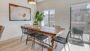 Uma área de refeições com mesa de madeira, cadeiras de metal preto, luminária pendente e uma grande janela com vista para prédios.