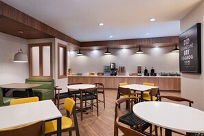 Um café moderno com mesas e cadeiras arredondadas, uma área de cozinha e uma parede com uma frase motivacional.