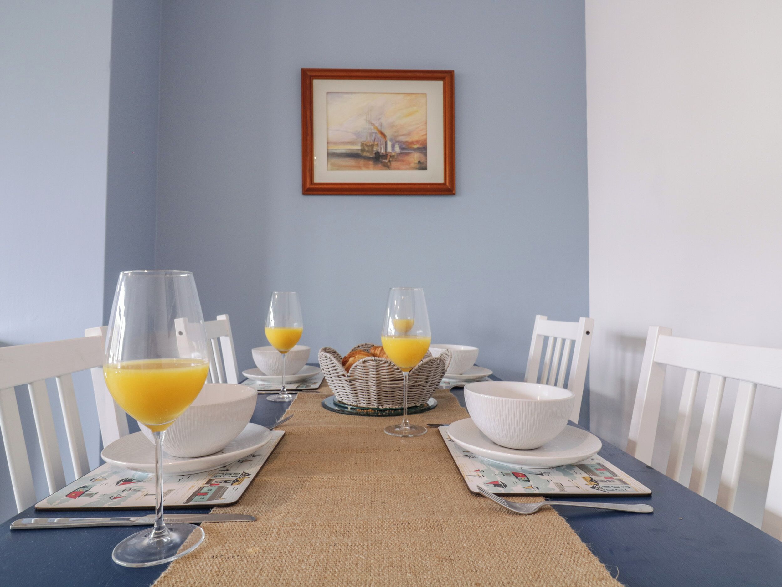 A dining table set with white plates, bowls, and glasses of orange juice, with a wicker basket and a framed painting on the wall.