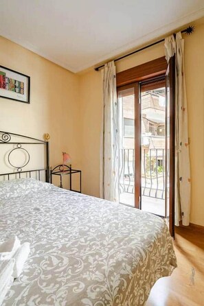 Quarto com cama, mesa de cabeceira, janela com cortinas e vista para o exterior.
