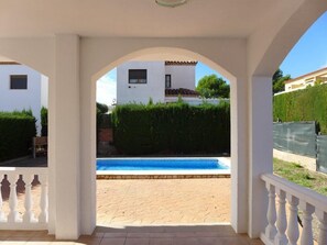 A covered outdoor area with a pool, white walls, and a view of a building with a tiled roof.