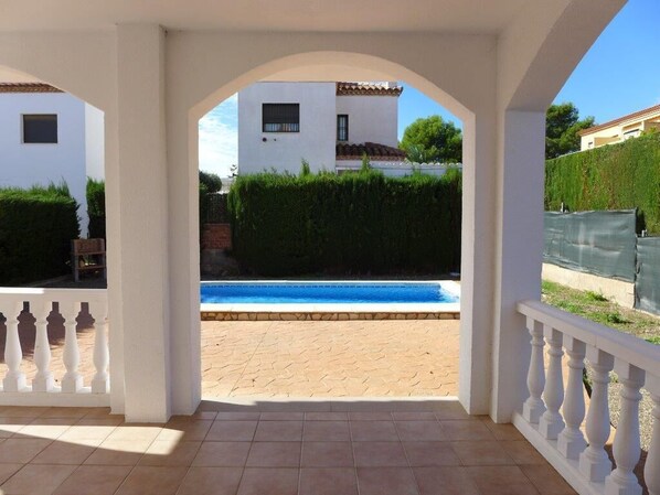 A covered outdoor area with a view of a swimming pool, a white building, and a hedge.