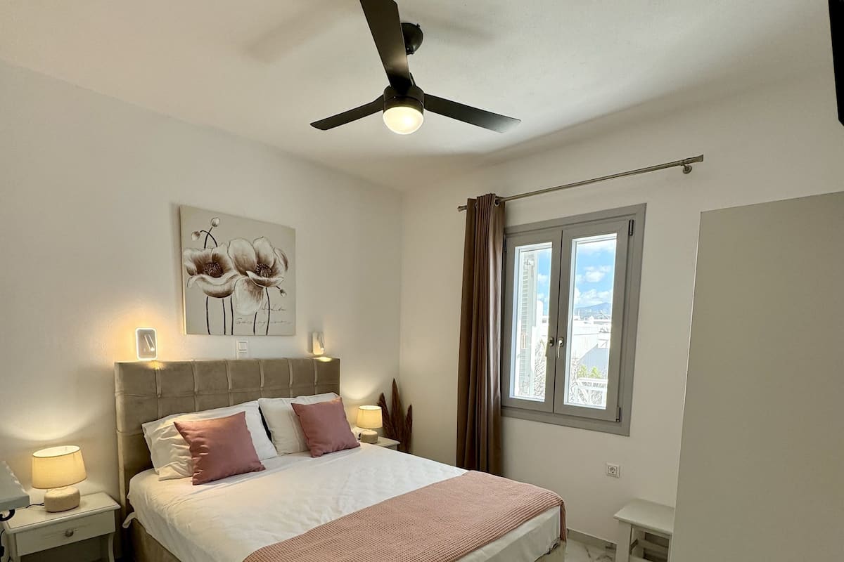 Une chambre à coucher comprenant un lit, des lampes de chevet, un ventilateur de plafond, une fenêtre avec des rideaux et une œuvre d’art accrochée au mur.
