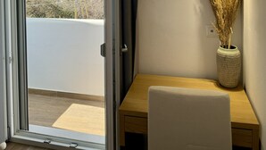 A room with a sliding glass door, a wooden desk, a white chair, a vase with dried plants, and a hanging pendant light. - Sunshine Bay Suites (Naxos)