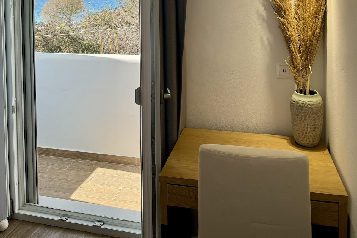 Une pièce dotée d’une porte-fenêtre coulissante, d’un bureau en bois, d’une chaise blanche, d’un vase contenant des plantes séchées et d’un luminaire suspendu.
