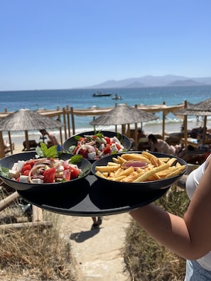 A tray with a salad and fries, held by a person at a beach with umbrellas and a view of the sea. - Sunshine Bay Suites (Naxos)