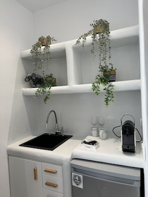 A kitchen with a black sink, white cabinets, and wooden handles. There are hanging plants and a coffee machine on the countertop. - Sunshine Bay Suites (Naxos)