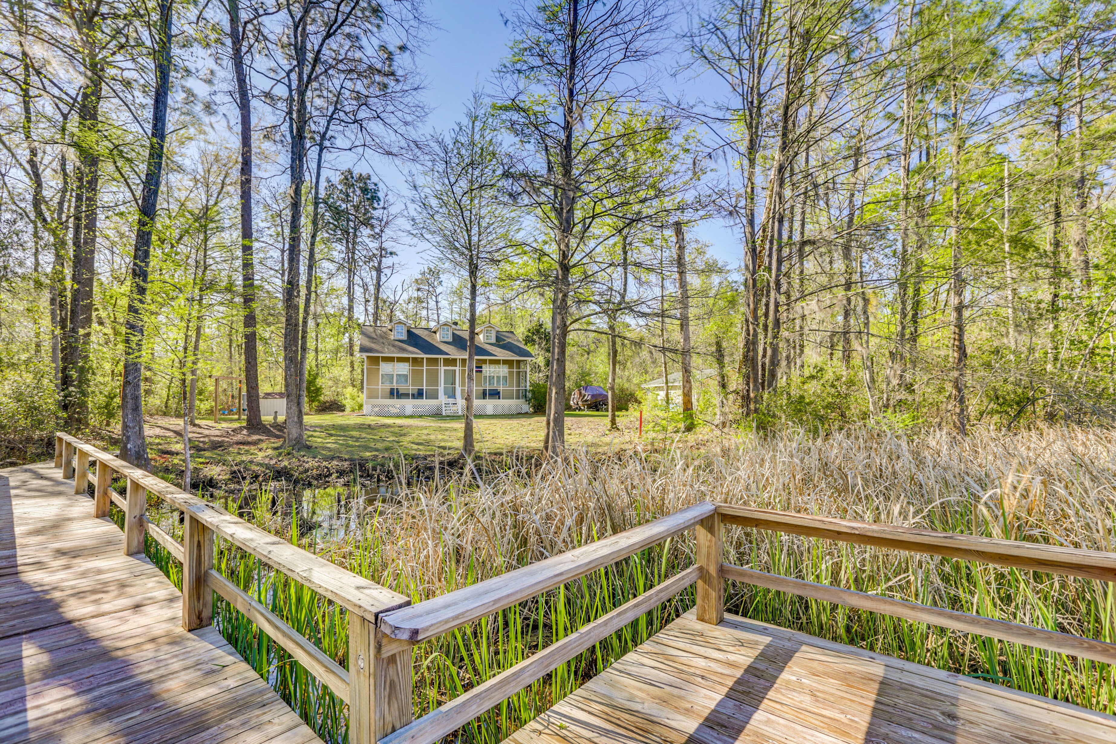 A wooden deck leading to a house surrounded by tall trees and a pond.