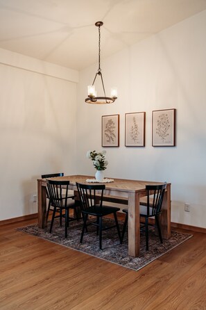Un coin repas comprenant une table en bois, quatre chaises, un luminaire suspendu et trois illustrations botaniques encadrées accrochées au mur.