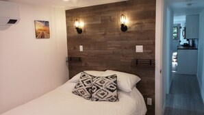 A bedroom with a wooden headboard, two wall-mounted lights, a bed with patterned pillows, a picture on the wall, and a hallway leading to a kitchen. - Luxury Caboose in Rhododendron Forest (Agassiz)
