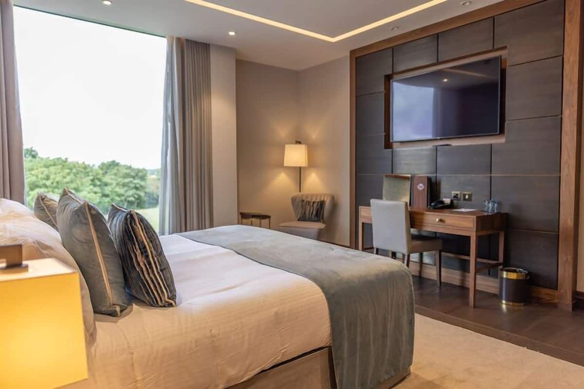 A modern hotel room with a large bed, a desk, a chair, a TV mounted on the wall, and a window with curtains.